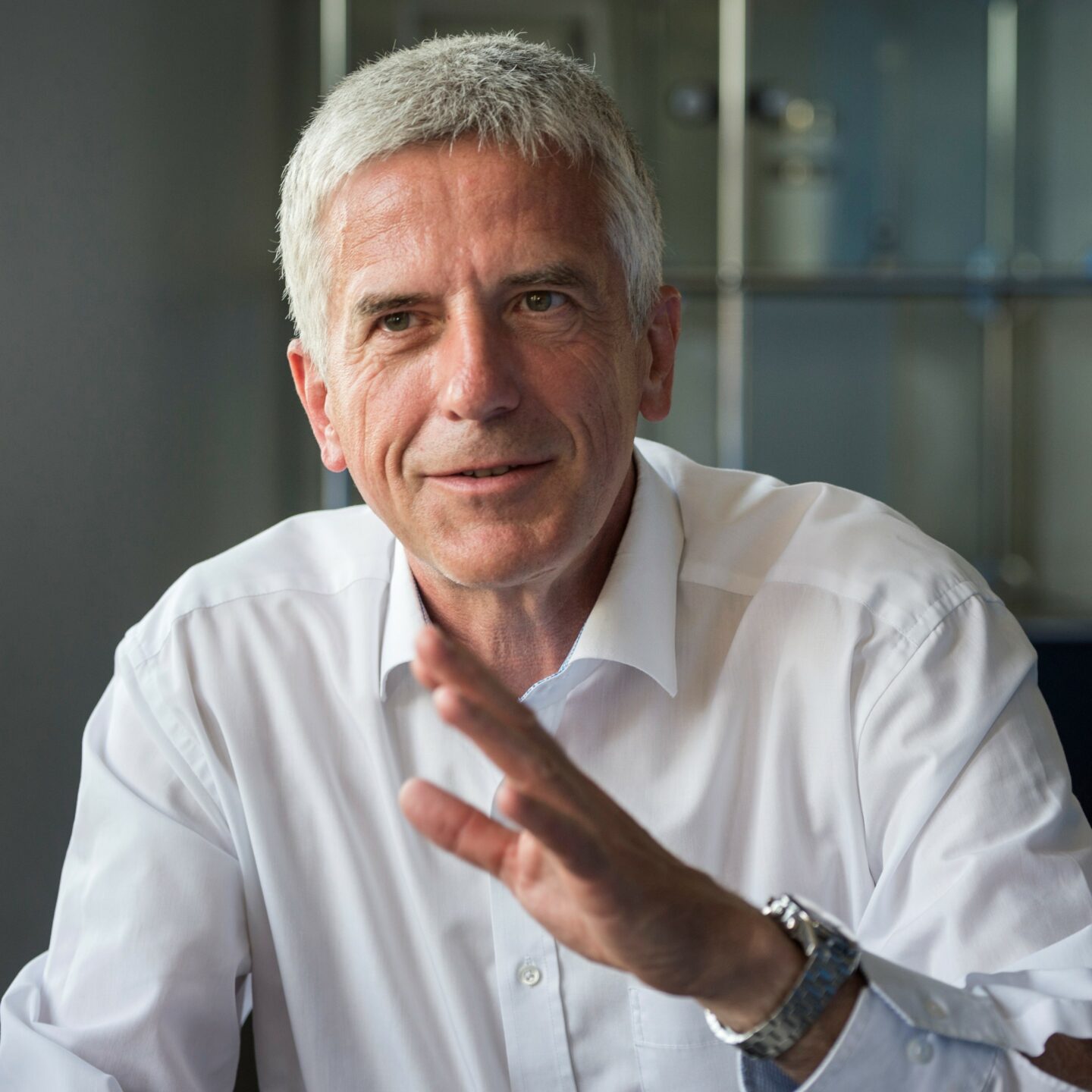 White-haired man in white shirt pictured explaining something with a hand gesture.