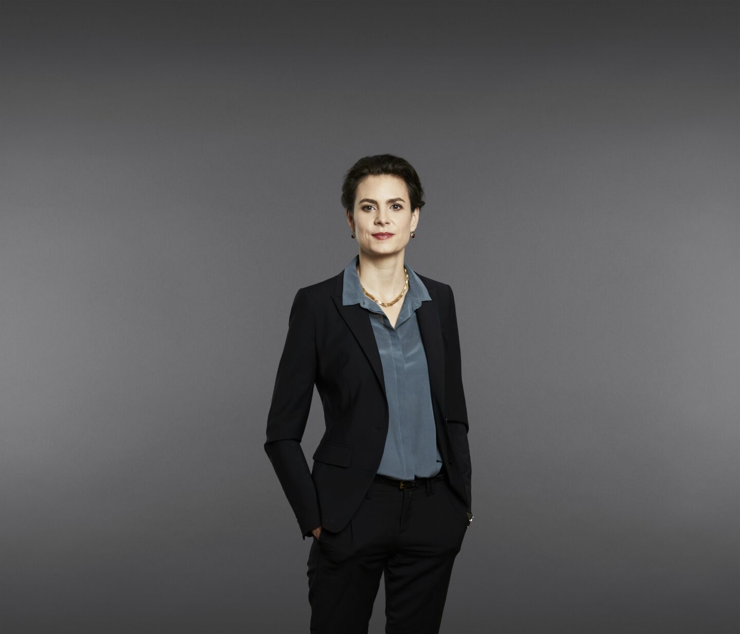 Slim woman in black, smart jacket and pants, with one hand in her pocket.