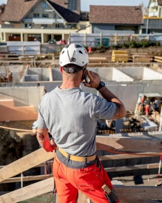 A foreman on the phone, from behind, wearing a safety helmet and watching over a construction site.
