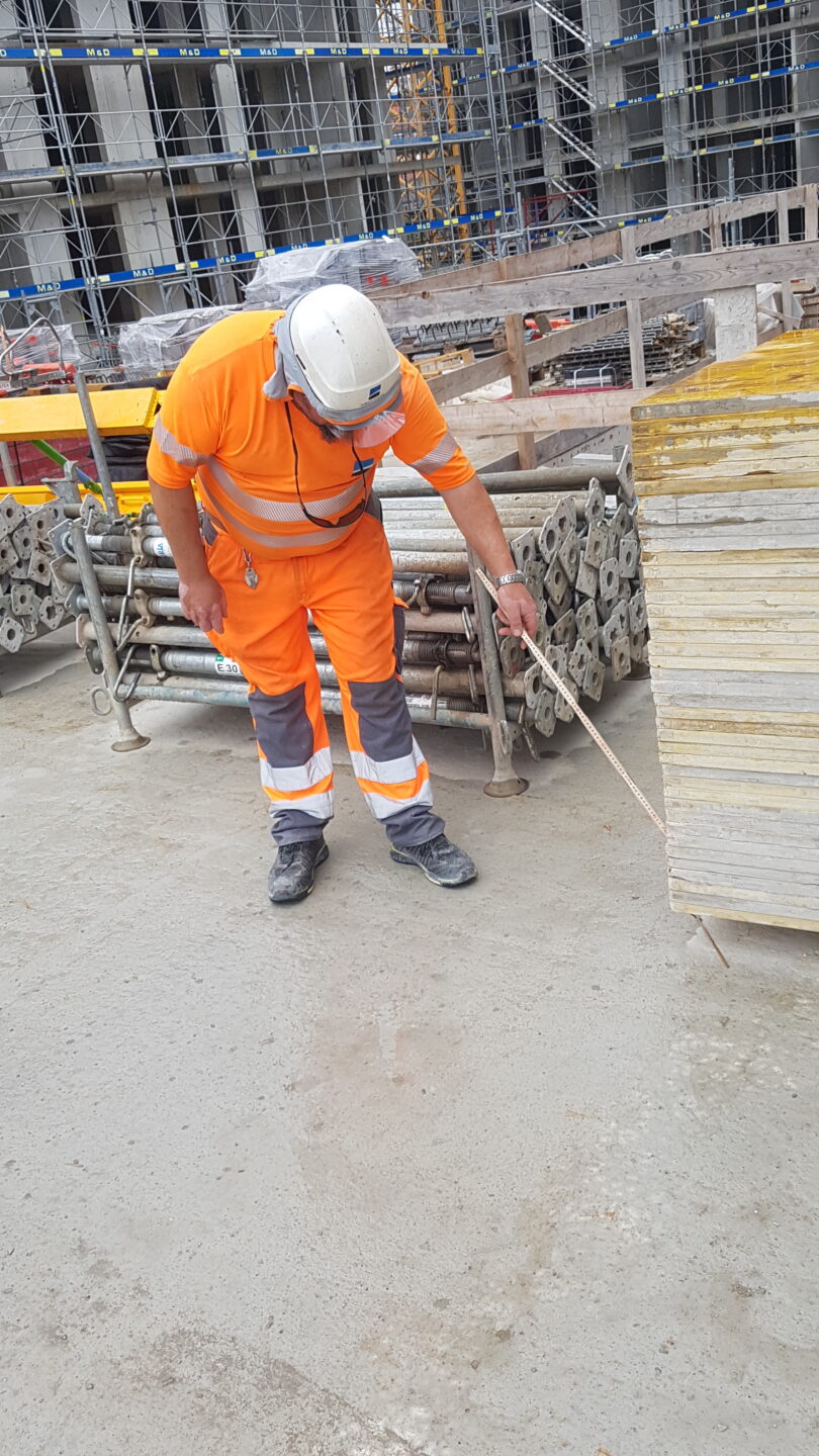 Foreman with front visor and cloth neck protection slightly leaning to measuring a pile.